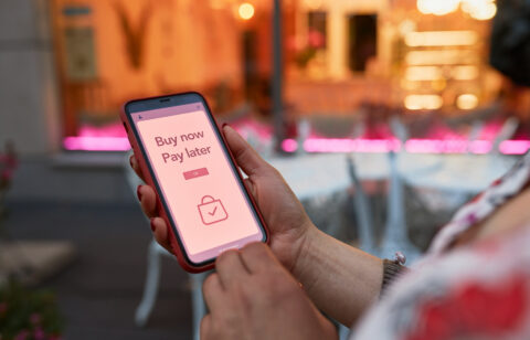 Person holding a smartphone displaying a "Buy now, pay later" message on a shopping app screen, with a blurred outdoor café background.