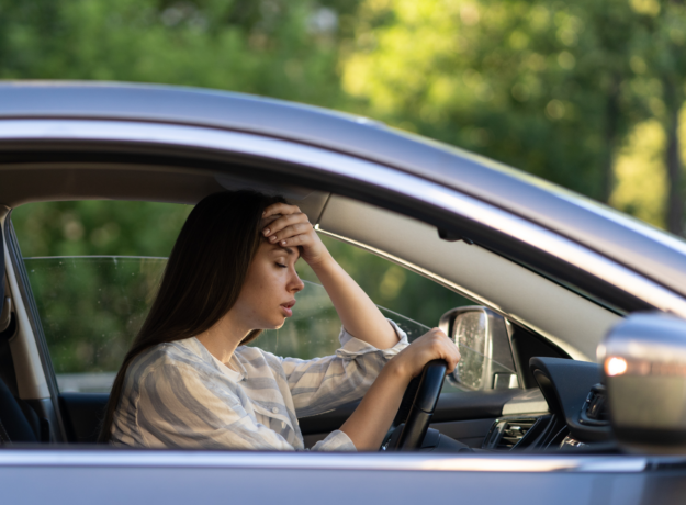 A woman sitting in a car with her hand on her forehead, looking stressed and overwhelmed.