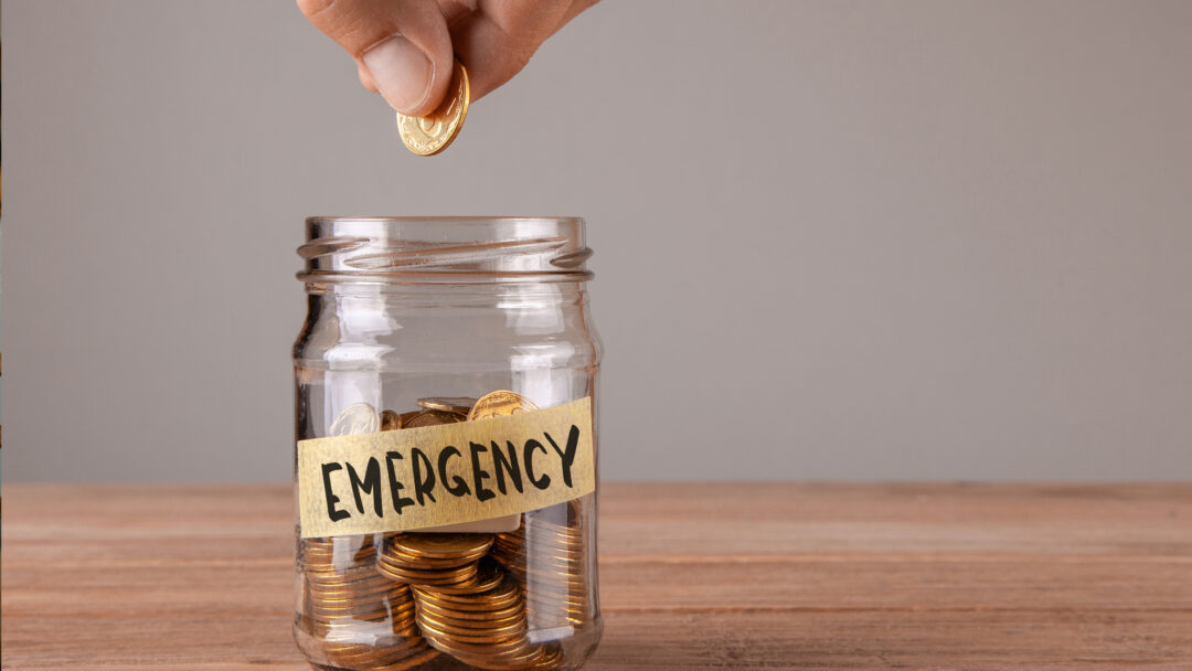 Hand placing a coin into a glass jar labeled “Emergency,” symbolizing saving money for unexpected expenses.