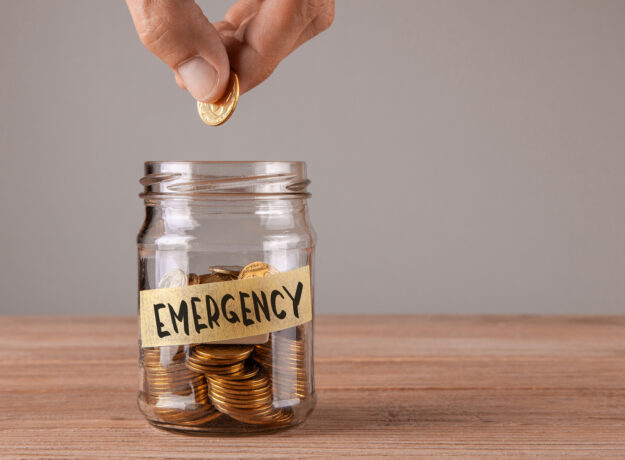 Hand placing a coin into a glass jar labeled “Emergency,” symbolizing saving money for unexpected expenses.