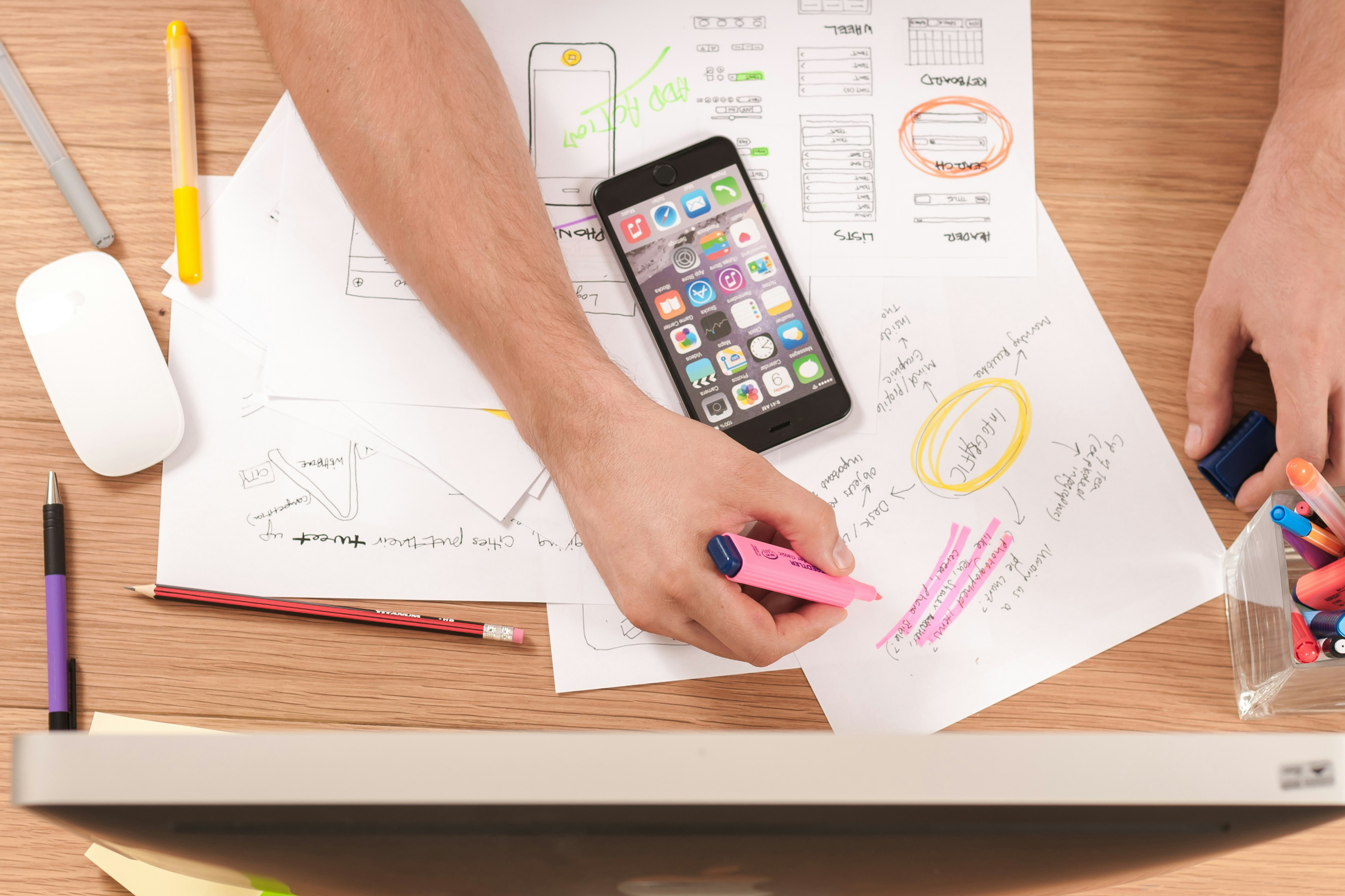 Person highlighting notes on wireframe sketches at a cluttered desk with a smartphone and colorful pens.