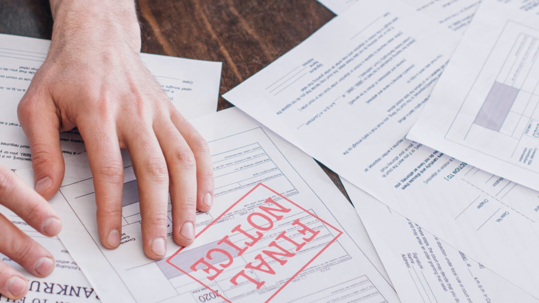 A person's hand resting on a stack of financial documents, with one paper prominently stamped “FINAL NOTICE” in red.
