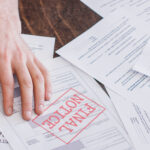 A person's hand resting on a stack of financial documents, with one paper prominently stamped “FINAL NOTICE” in red.