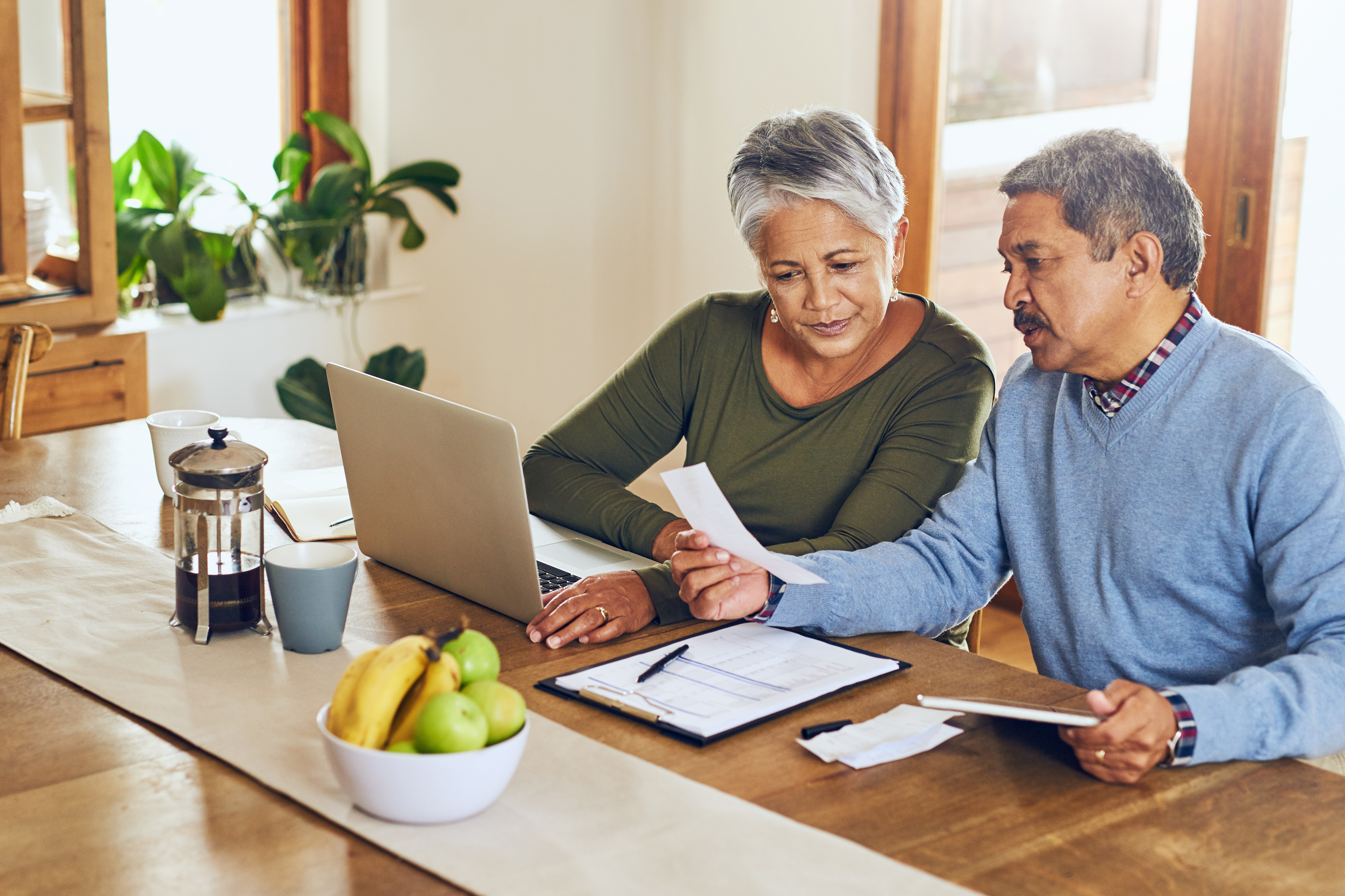 Older couple sitting at a kitchen table reviewing financial documents together with a laptop and tablet.