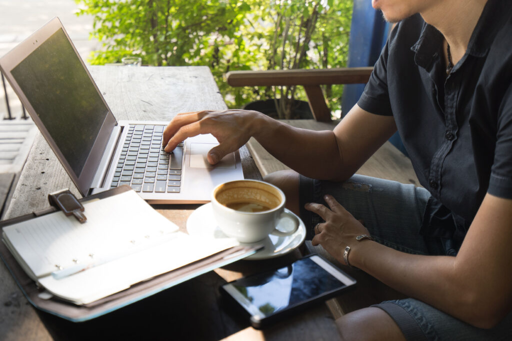 Person working on a laptop at an outdoor café table with a cup of coffee, a smartphone, and a notebook nearby.