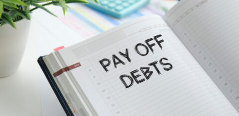 Close-up of an open planner with the handwritten words “Pay Off Debts” on a dated page, surrounded by office supplies.