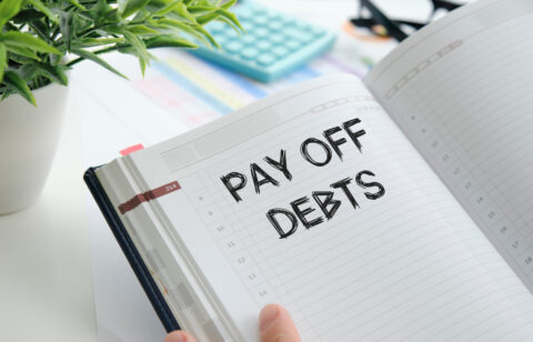 Close-up of an open planner with the handwritten words βPay Off Debtsβ on a dated page, surrounded by office supplies.