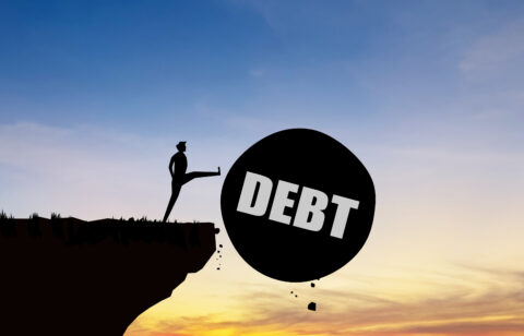 Silhouette of a person kicking a large ball labeled "DEBT" off a cliff at sunset.