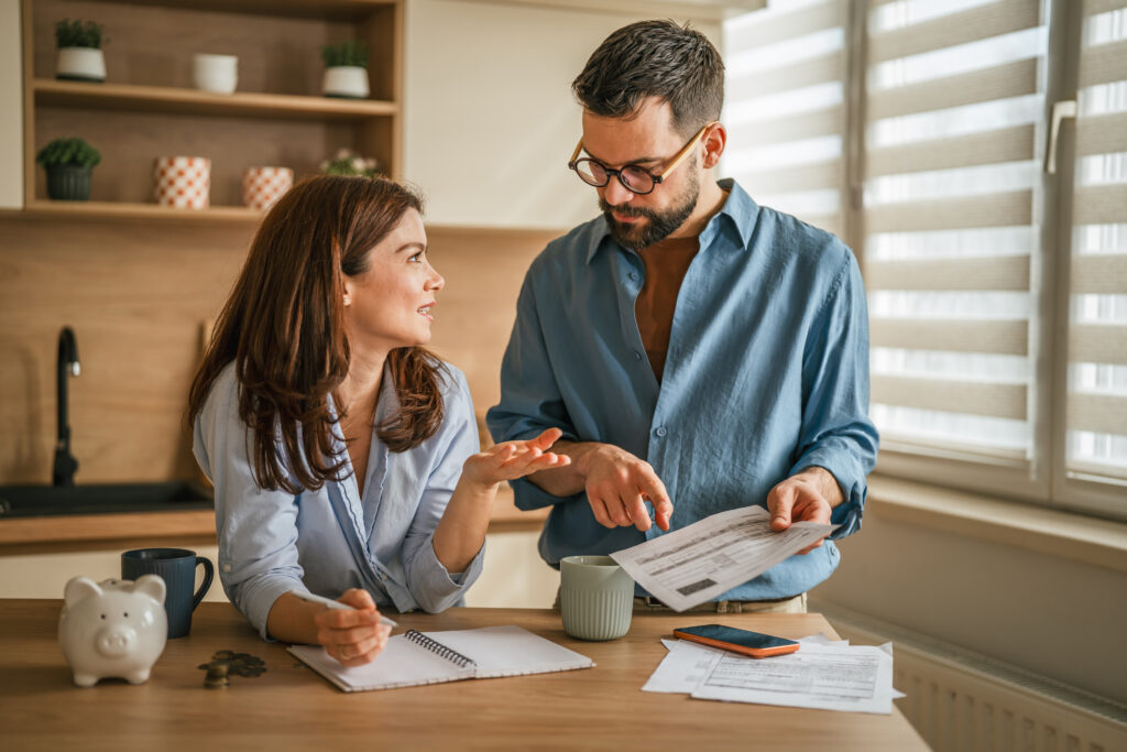 Couple reviewing household bills and budgeting together at a kitchen table with paperwork, coffee mugs, and a piggy bank nearby.