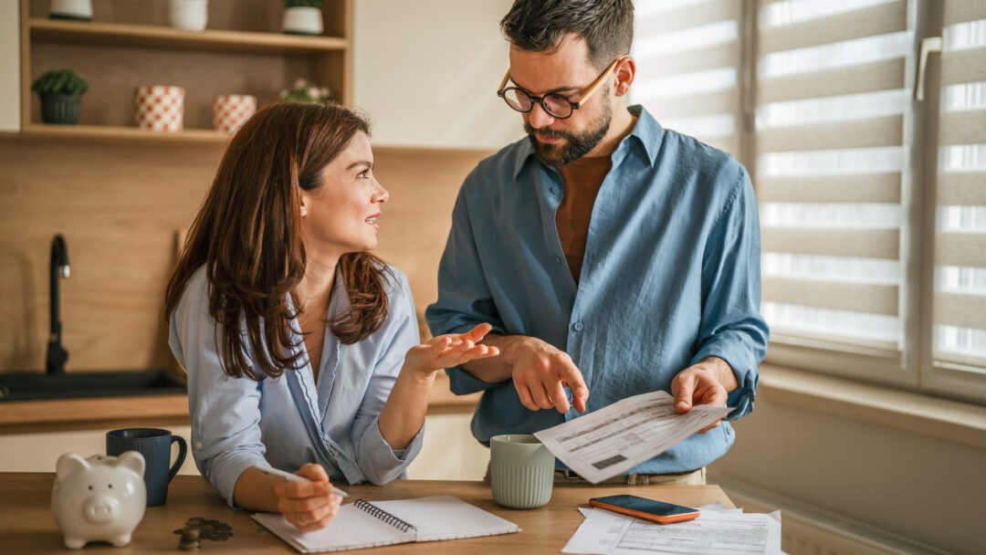 Couple reviewing household bills and budgeting together at a kitchen table with paperwork, coffee mugs, and a piggy bank nearby.