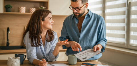 Couple reviewing household bills and budgeting together at a kitchen table with paperwork, coffee mugs, and a piggy bank nearby.