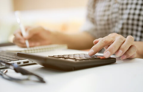 Close-up of a person using a calculator while taking notes next to a laptop on a desk.