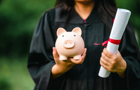 Graduate in a cap and gown holding a piggy bank and a diploma tied with a red ribbon