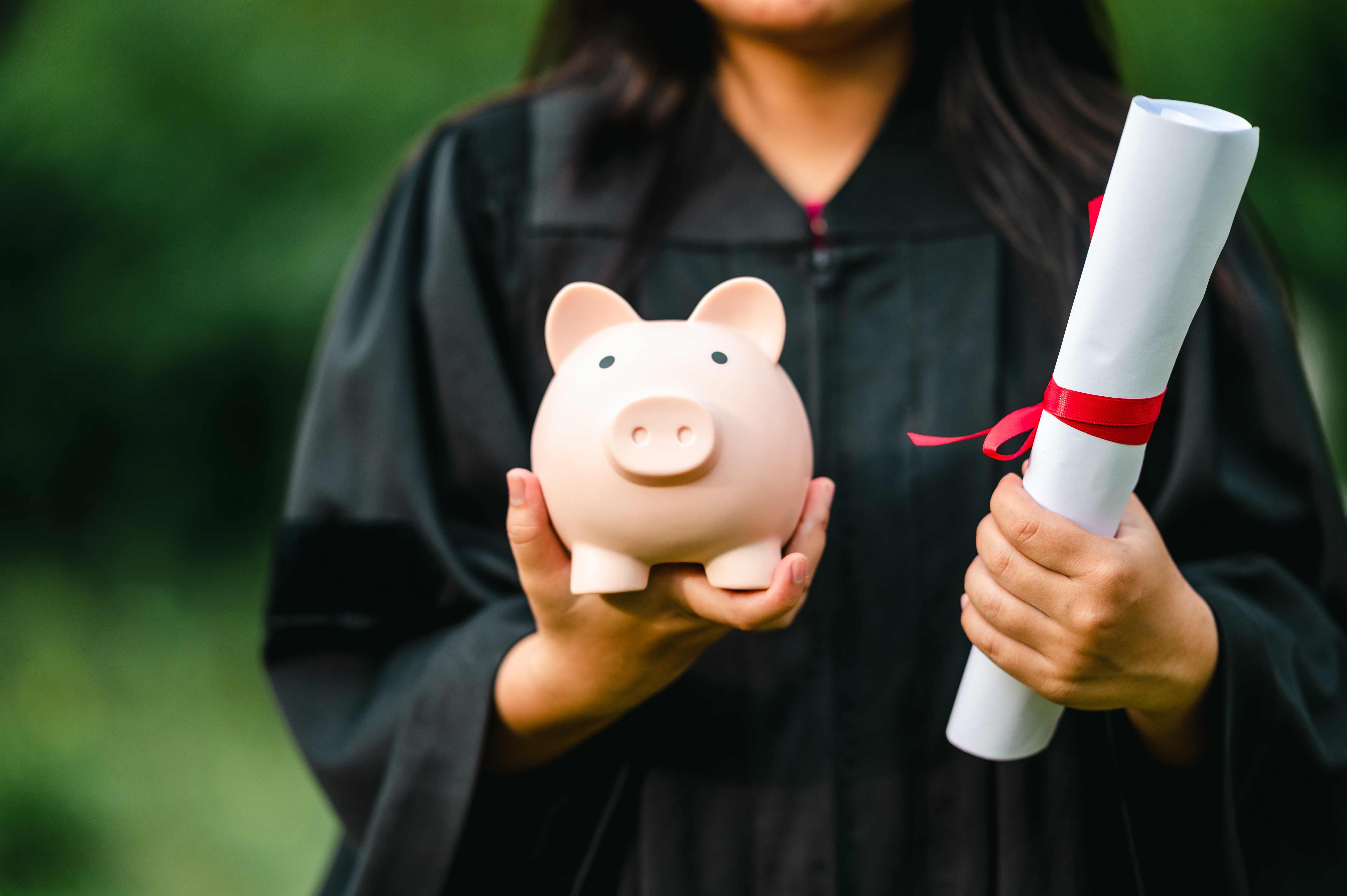 Graduate in a cap and gown holding a piggy bank and a diploma tied with a red ribbon