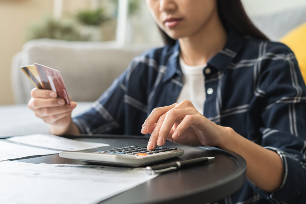 Close-up of a woman using a calculator while holding credit cards, with bills and paperwork spread out on a table.