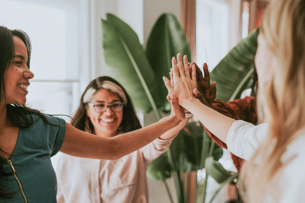 A group of smiling women standing in a circle, giving a group high-five in a bright indoor space with plants in the background.