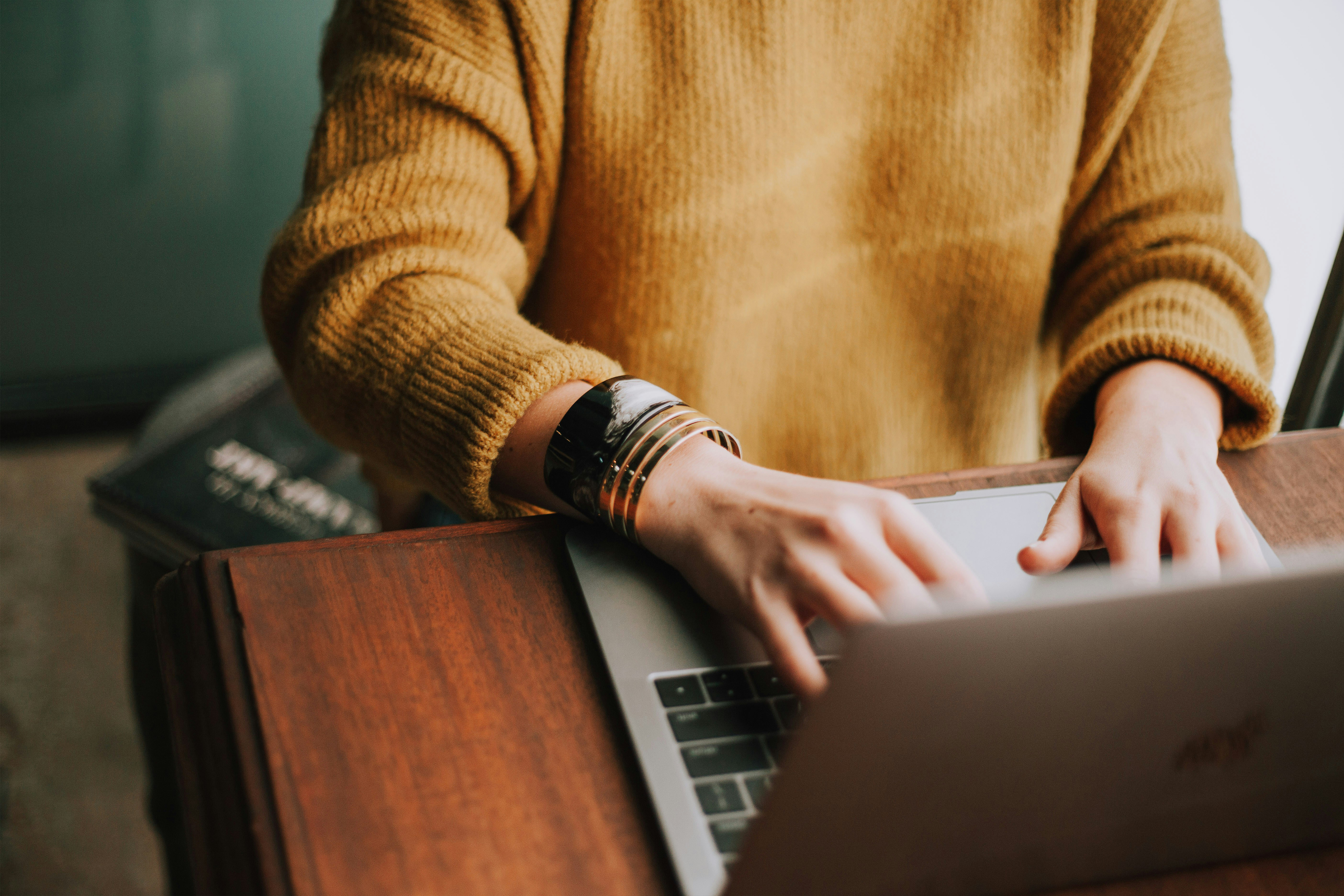 Person in a yellow sweater typing on a laptop at a wooden desk, with a bracelet on their wrist.