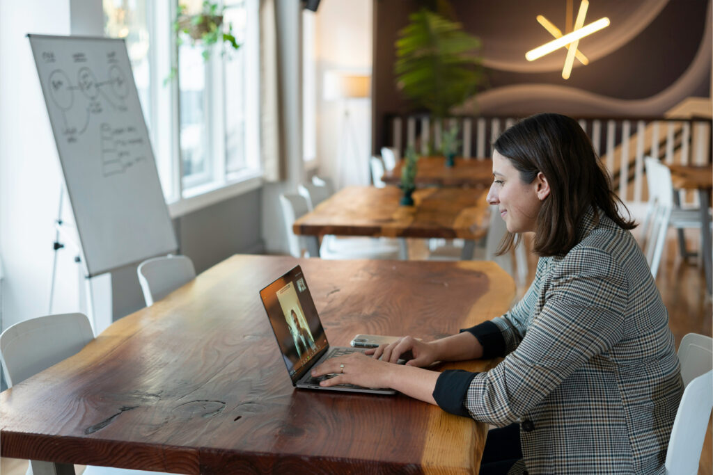 Professional woman in a plaid blazer sitting at a wooden table, smiling during a video call on her laptop in a modern, well-lit workspace.