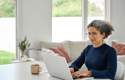 Middle-aged woman working on a laptop at home, smiling slightly while seated at a bright, modern table with a coffee mug nearby.