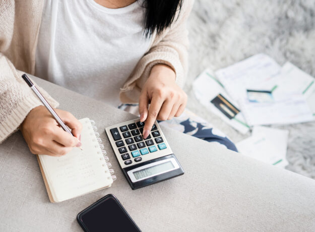 Woman calculating expenses with a large calculator and taking notes in a notebook, surrounded by bills and a smartphone.