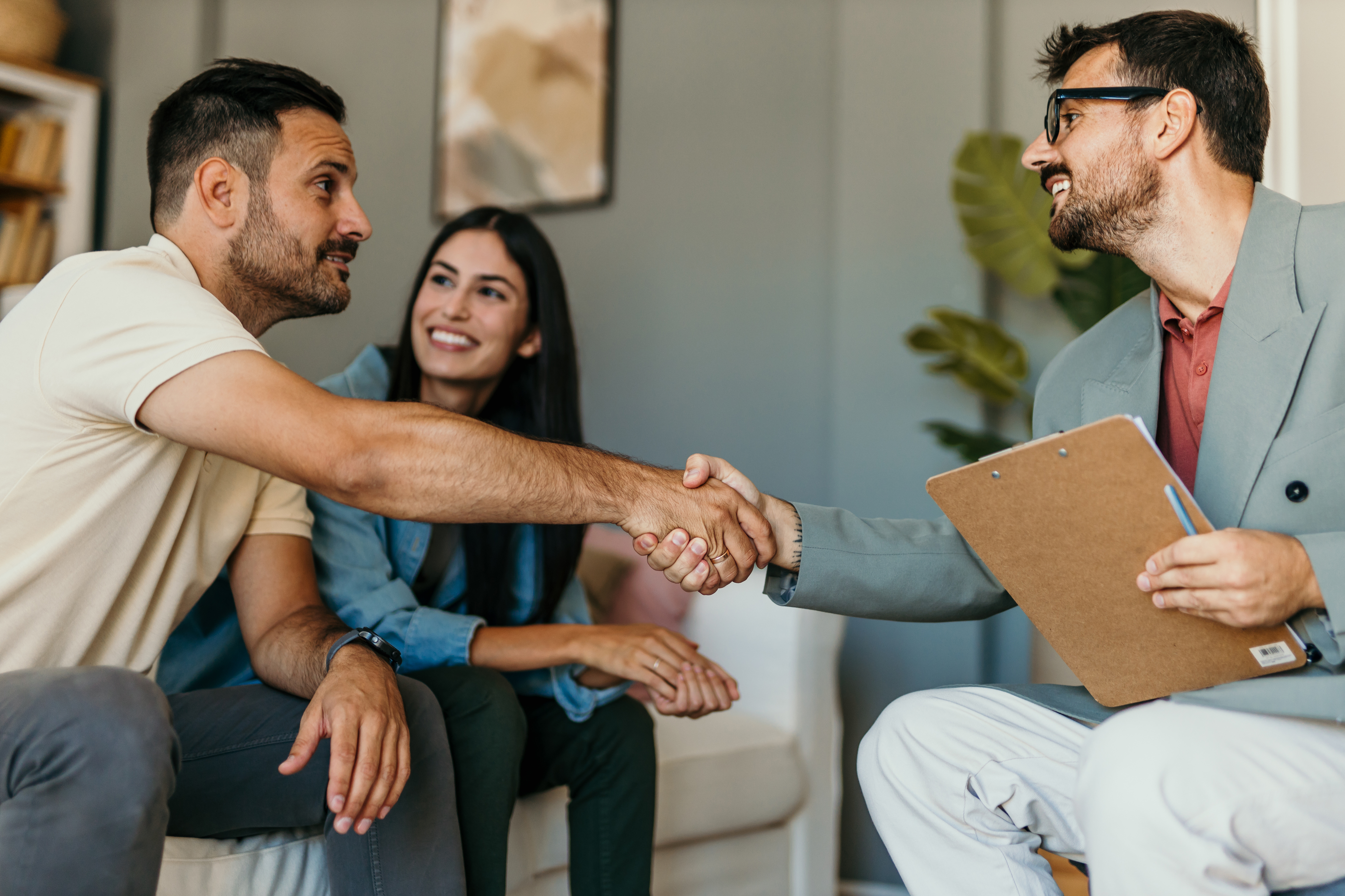 Smiling couple meeting with a professional holding a clipboard, as the man and the advisor shake hands in a friendly office setting.
