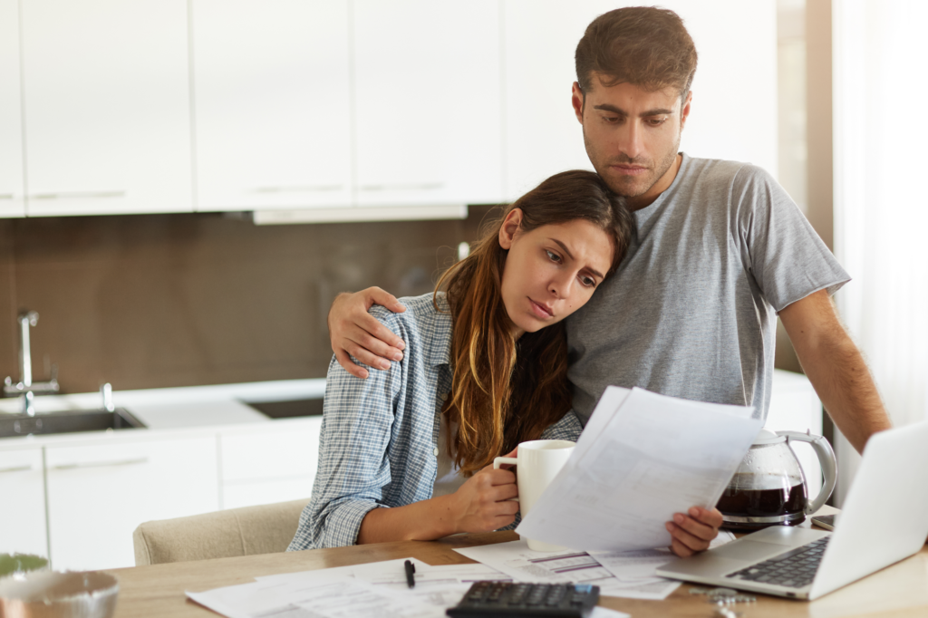 A worried couple sits at a kitchen table reviewing bills and financial documents, with a laptop, coffee pot, and calculator in front of them.