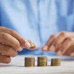 Close-up of a person in a blue shirt stacking coins into small piles on a table, representing saving or budgeting money.