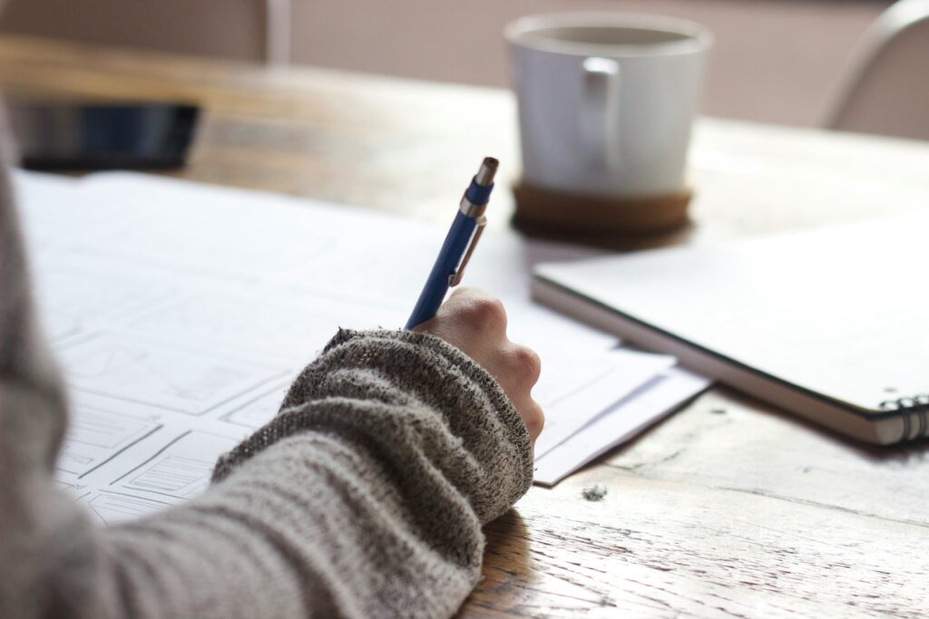 Close-up of a person writing on paper at a wooden desk with a notebook and coffee mug nearby.