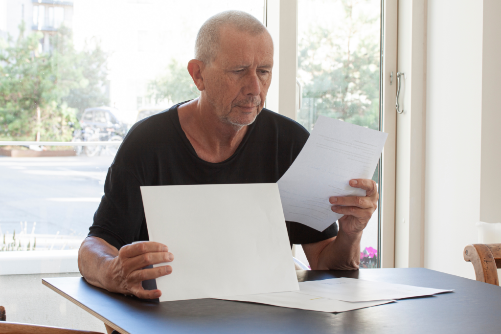 Older man sitting at a table reading documents in a brightly lit room.