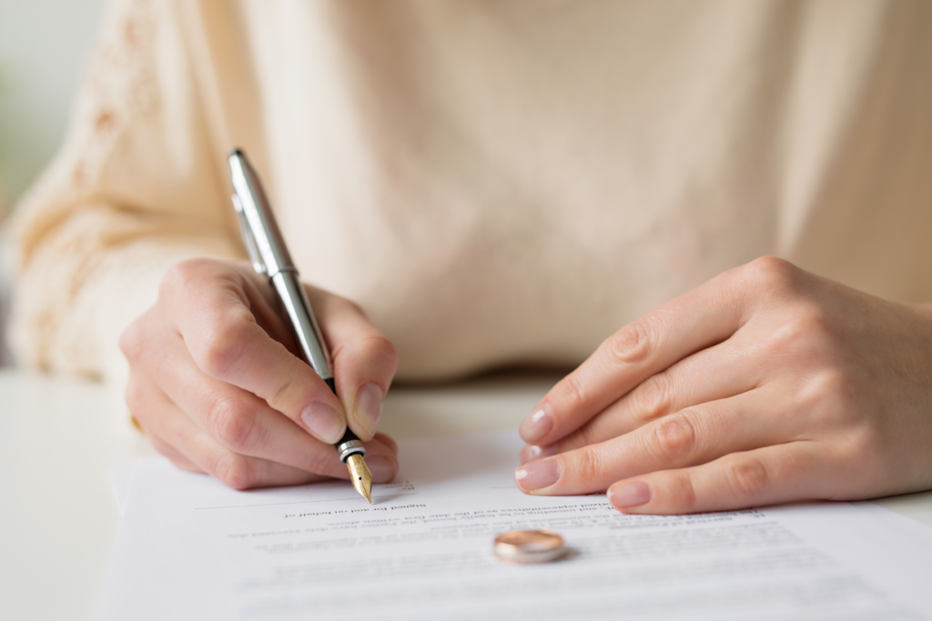 Close-up of a woman signing a legal document with wedding rings placed on the paper.