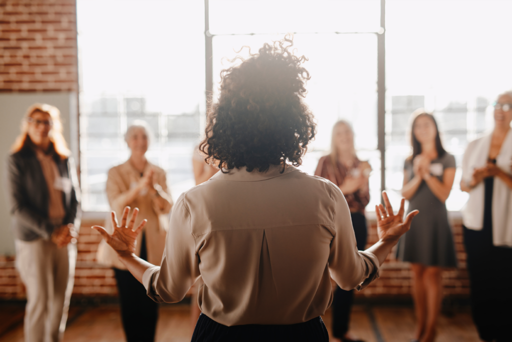 Back view of a woman speaking to a group of people in a bright room, with several women standing and clapping.
