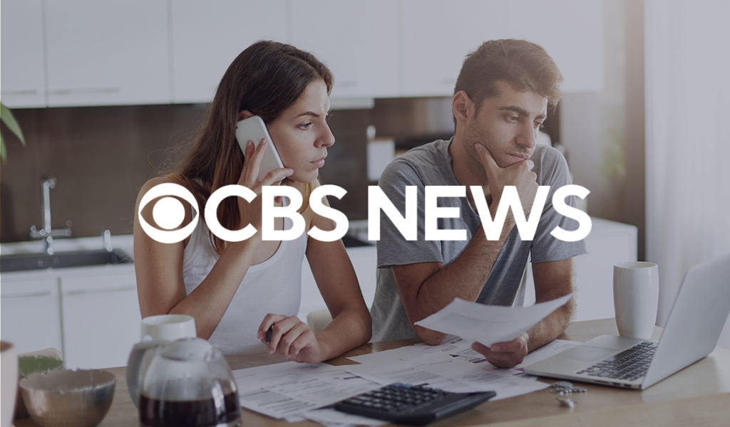 Man and woman reviewing bills at a kitchen table with a laptop and calculator, appearing concerned. CBS News logo overlaid.