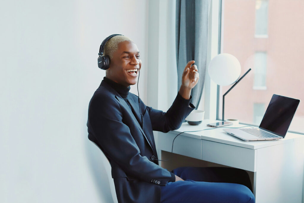 Smiling man wearing headphones sitting at a desk with a laptop, engaged in a virtual conversation.