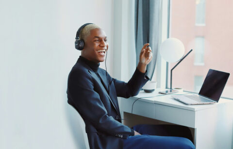 Smiling man wearing headphones sitting at a desk with a laptop, engaged in a virtual conversation.