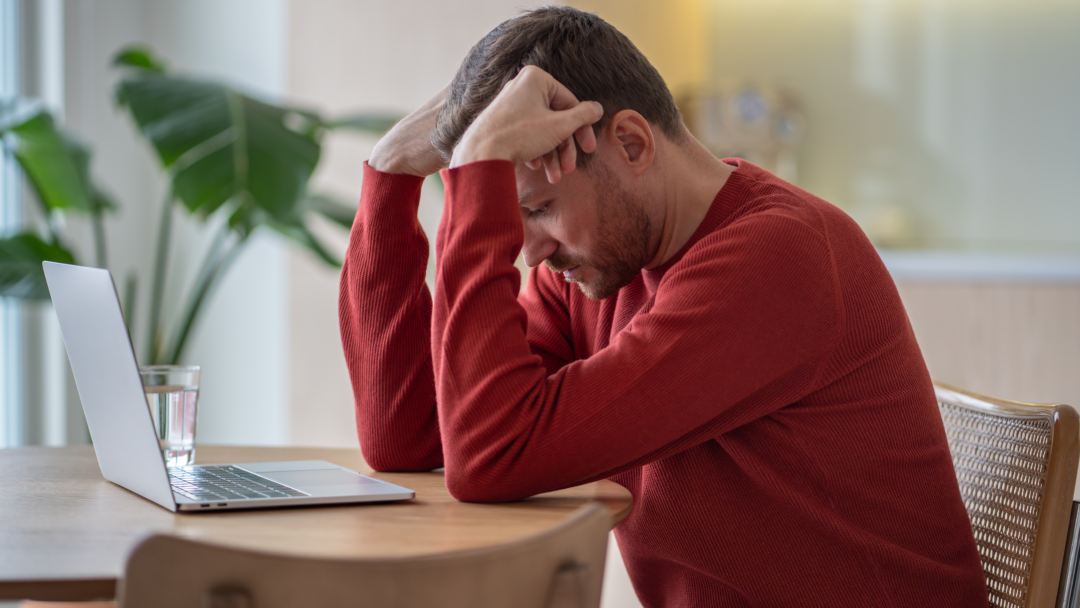 Man in a red sweater sitting at a table with his head in his hands, looking stressed while using a laptop.