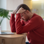 Man in a red sweater sitting at a table with his head in his hands, looking stressed while using a laptop.
