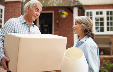 Older couple smiling at each other while carrying moving boxes and a lamp outside a brick home.