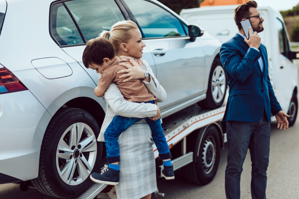 Woman holding a child beside a car being towed, while a man in a suit makes a phone call.