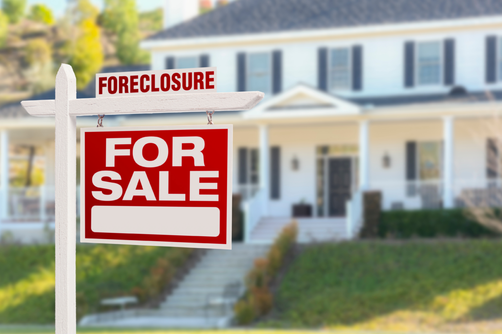 Red "For Sale" sign with "Foreclosure" label in front of a large, white house.
