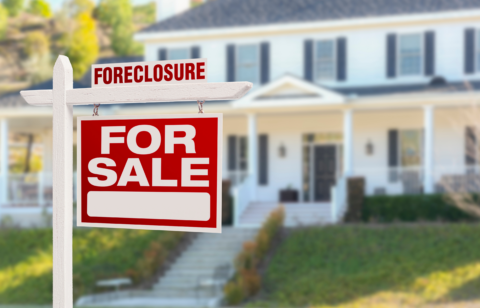 Red "For Sale" sign with "Foreclosure" label in front of a large, white house.