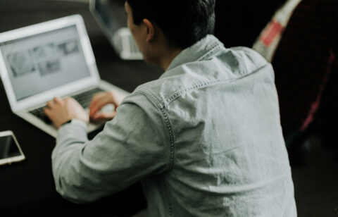 Person working on a laptop at a desk, viewed from behind, with a smartphone placed nearby.