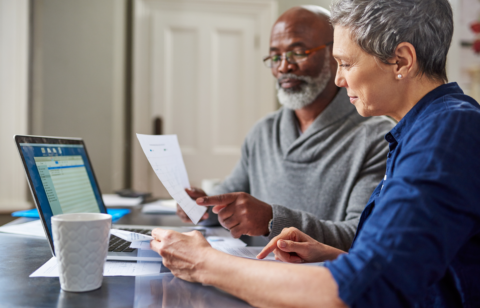 Older couple reviewing documents together at a kitchen table, with a laptop open and papers spread out in front of them.