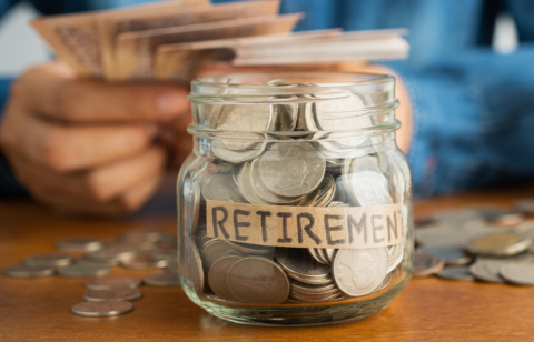 Glass jar labeled 'Retirement' filled with coins, with a person in the background holding currency notes.
