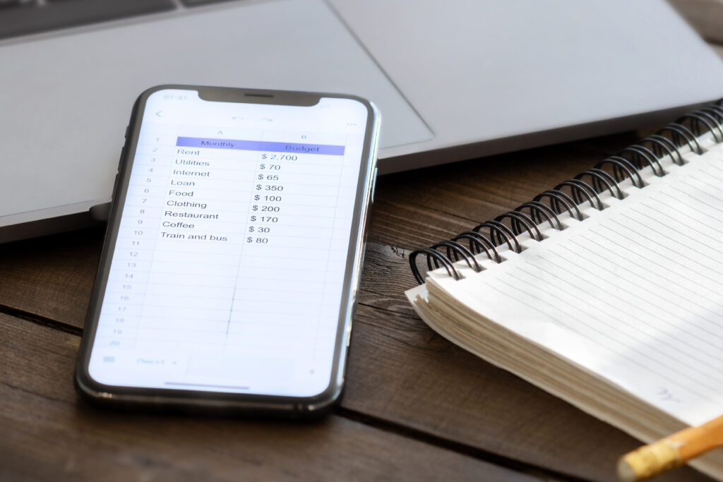 Smartphone displaying a personal monthly budget spreadsheet next to a spiral notebook and laptop on a wooden desk.