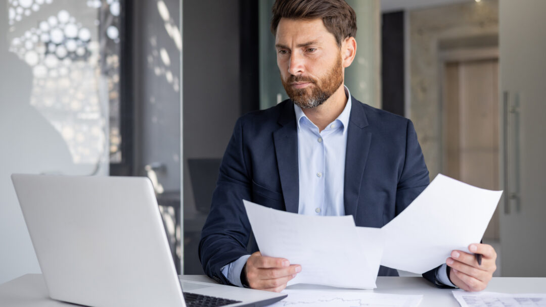 A serious young man businessman works in the office at a desk, looks at the screen of a laptop, holds documents in his hands, compares data, makes statistics and analysis