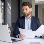 A serious young man businessman works in the office at a desk, looks at the screen of a laptop, holds documents in his hands, compares data, makes statistics and analysis