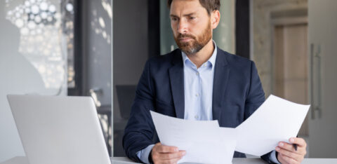 A serious young man businessman works in the office at a desk, looks at the screen of a laptop, holds documents in his hands, compares data, makes statistics and analysis