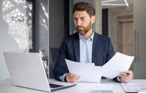 A serious young man businessman works in the office at a desk, looks at the screen of a laptop, holds documents in his hands, compares data, makes statistics and analysis