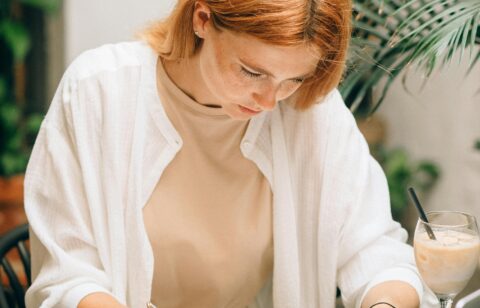 Woman reviewing documents and writing on paper at a desk with a laptop, calculator, and iced coffee