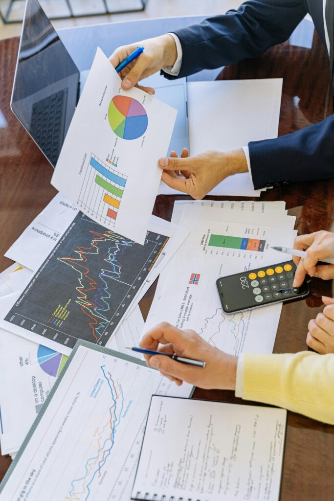 Two professionals review printed charts and graphs at a meeting table, using a calculator and laptop to analyze financial or marketing data.